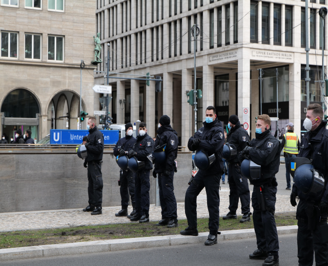 Polizisten in schwarzen Uniformen und Masken stehen vor einem Berliner Gebäude mit Glasfenstern und Säulen, einige halten Helme, mit Laternen, Verkehrszeichen, Schildern mit Text, einer Statue und anderen Menschen im Hintergrund.