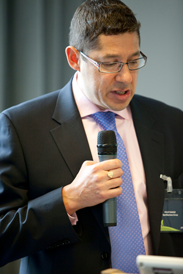 Ein Mann in Anzug, Hemd und Krawatte mit Brille hält ein Mikrofon und spricht, mit einem weißen Abzeichen am Hemd und einem Vorhang im Hintergrund.