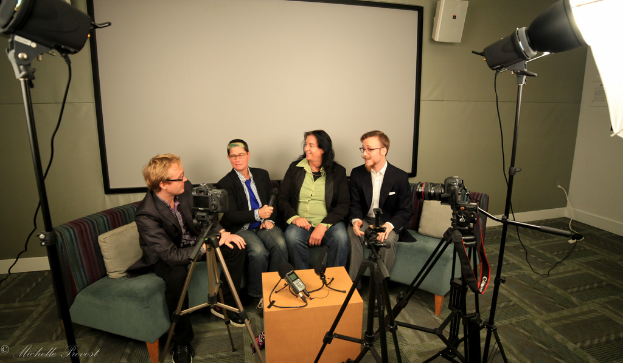Eine Gruppe von drei Personen sitzt auf einem Sofa und gibt ein Interview. Eine Person in einem schwarzen Mantel und Hose ist links zu sehen, eine Person in einem Mantel und blauer Krawatte hält in der Mitte ein Mikrofon, und eine Person rechts lächelt. Ein Mikrofonrekorder steht in der Mitte auf dem Tisch, Kameras sind auf der rechten und linken Seite zu sehen, sowie große Scheinwerfer, und ein Projektionsschirm im Hintergrund.