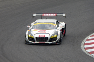 Ein leuchtend rotes und weißes Audi R8 LMS GT3 Rennfahrzeug mit dem Audi-Logo auf der Seite, gefahren von einer Person in einem schwarzen Rennanzug und Helm, die um eine Rennstrecke fährt.