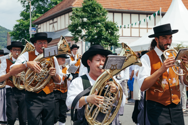 Eine Gruppe von Männern in traditioneller bayerischer Tracht marschiert die Straße entlang, spielt Brass-Instrumente und hält Bücher in den Händen, mit Zelten, Gebäuden, Bäumen, einem Pfahl, einem Schild, einem Berg und einem klaren blauen Himmel im Hintergrund.
