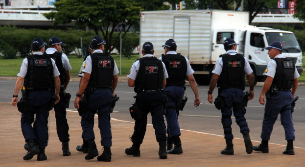 Eine Gruppe von Beamten in Uniform, darunter Mützen, Hemden, Schutzwesten, Uhren, Hosen und Schuhe, geht auf einer Straße. Rechts daneben parkt ein Lastwagen in der Nähe eines Zauns, mit Gebäuden, Bäumen, Pflanzen, Pfählen und Bannern im Hintergrund und einer Brücke oben rechts.