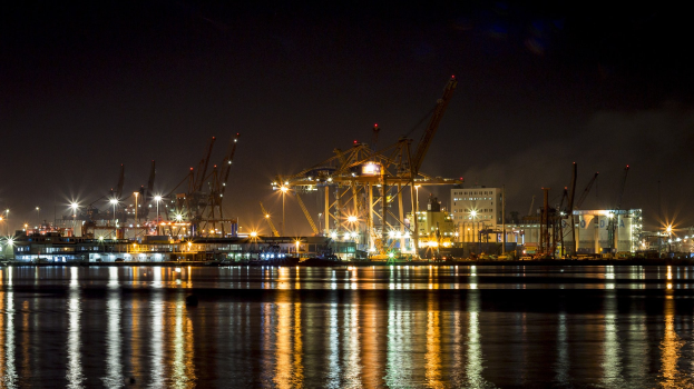 Großes Container-Schiff nachts am Kai, beleuchtet von umliegenden Gebäuden und Kränen, mit dunklem Himmel.