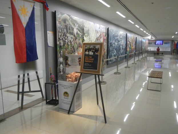 Langer Flur mit einer philippinischen Flagge an der Wand, Informationsschilder, Ribbon-Ständer, Bänke, Deckenleuchten und ein Bildschirm mit Museums-Informationen im Hintergrund.