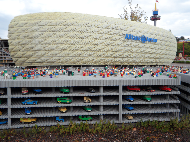 Eine große Lego-Struktur, die dem Allianz Stadion in München, Deutschland, ähnelt, mit einer Gruppe von Menschen davor, umgeben von Bäumen, Pfählen, einem Turm und Grünfläche, unter einem bewölkten Himmel.