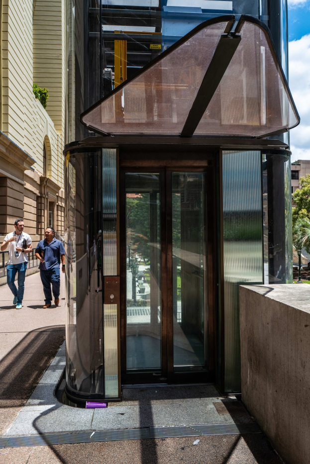 Glasaufzug umgeben von Fußgängern in einer städtischen Straße, mit einer Wand auf der rechten Seite, Gebäuden, Bäumen und einem bewölkten Himmel im Hintergrund.