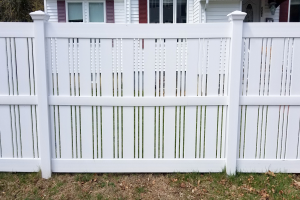 Ein weißer Vinyl-Privacy-Zaun steht vor einem Haus mit Fenstern, umgeben von Gras und trockenen Blättern, mit Bäumen im Hintergrund.