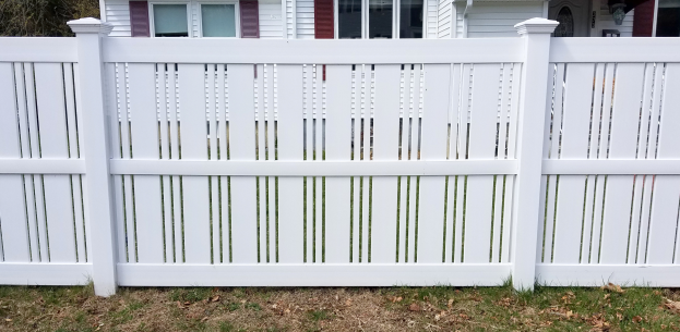 Ein weißer Vinyl-Privacy-Zaun steht vor einem Haus mit Fenstern, umgeben von Gras und trockenen Blättern, mit Bäumen im Hintergrund.