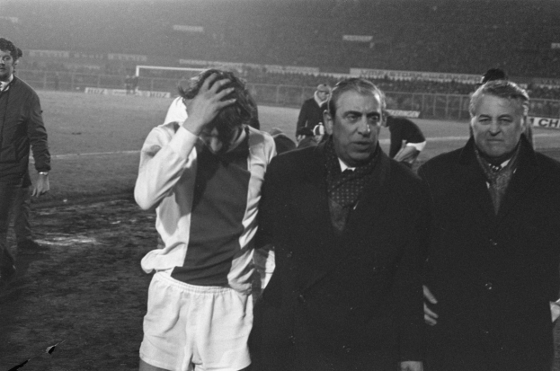 Schwarzes und weißes Bild einer Gruppe von Männern auf einem Fußballfeld mit einem Stadion im Hintergrund, die enttäuscht nach einem Sieg aussehen.