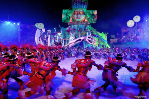 Eine Gruppe von Menschen in farbenfrohen Kostümen tanzt auf einer Bühne vor einem sitzenden Publikum, mit Lichtern, Pfählen und Ballons, und ein Bildschirm zeigt eine Person im Hintergrund, unter einem sichtbaren Himmel, bei der Rio-Karneval 2014.