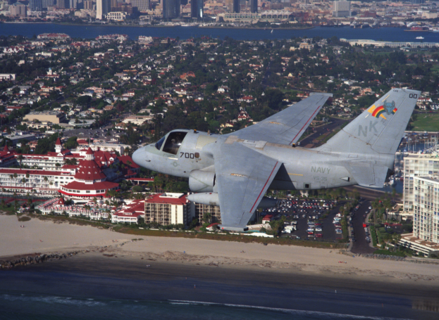 Ein Kampfflugzeug fliegt über einen Strand neben einer Stadt, mit Gebäuden, Bäumen, Fahrzeugen und Wasser im Hintergrund.