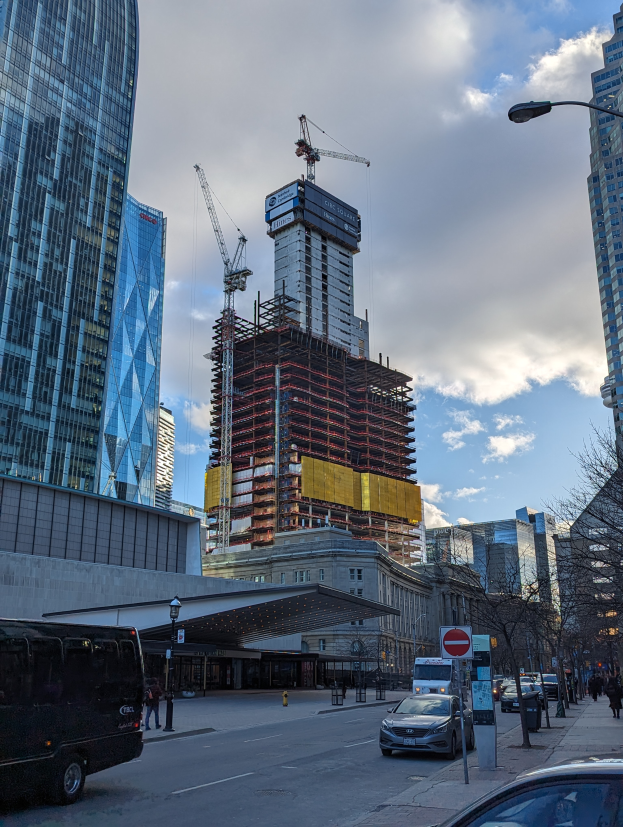Eine belebte Stadtstraße mit Fahrzeugen, Fußgängern, Schildern, Laternen und Bäumen, mit dem One World Trade Center im Hintergrund unter einem bewölkten Himmel.