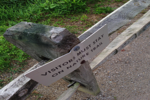Ein hölzerner Zaun mit einem Schild, auf dem "Besucher müssen auf dem Naturweg bleiben", umgeben von grünem Gras und kleinen Steinen.