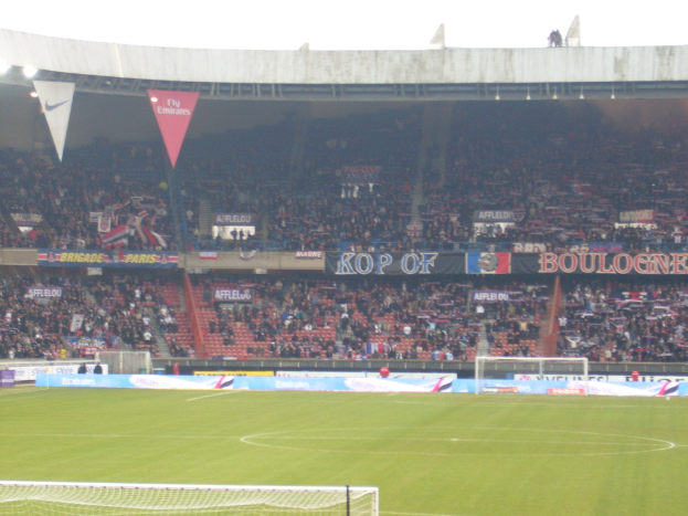 Ein grüner Stadionboden mit Torständen auf beiden Seiten, umgeben von sitzenden Zuschauern und geschmückt mit zahlreichen Werbetafeln, unter einem klaren Himmel.