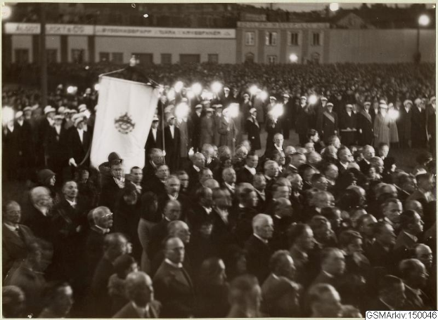 Schwarzes und weißes Foto einer großen Menschenmenge vor einem Gebäude, mit einer Person in der Mitte, die eine Fahne hält, sowie Masten und Lichter im Hintergrund und einem Text unten, der "Gustav Klimts Rede an der Berliner Mauer" lautet.
