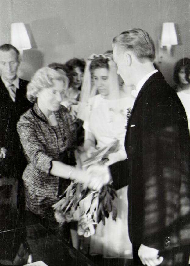 Ein Schwarz-Weiß-Foto eines Brautpaares beim Austausch der Eheringe, wobei die Braut in einem weißen Kleid und Schleier mit einem Strauß Blumen in der Hand zu sehen ist und der Bräutigam in einem Anzug und Krawatte steht, mit Wandlampen im Hintergrund.