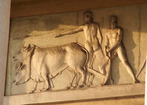 Steinwandrelief mit zwei Menschen, die mit Hilfe von zwei Kühen ein Feld pflügen.