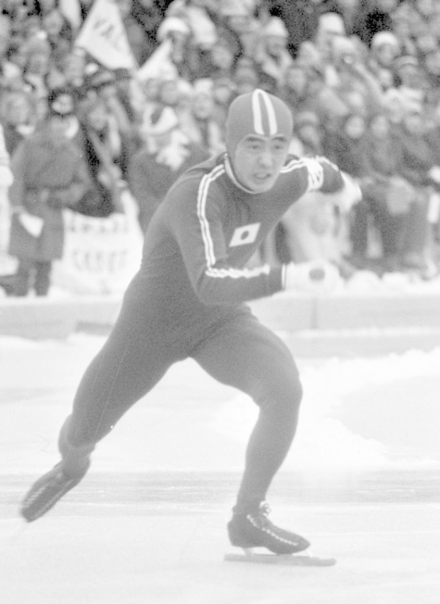 Ein Schwarz-Weiß-Foto eines Eisschnellläufers in Aktion auf dem Eis, der einen Helm und Eislaufschuhe trägt, mit ein paar Menschen im Hintergrund, die Flaggen halten.