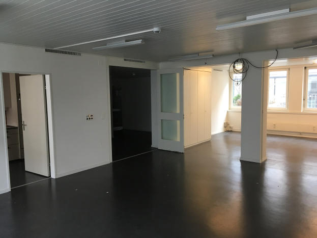 Leerer Büroraum mit schwarzen Böden, weißen Wänden, elektrischem Licht, Kabeln, Türen, Schränken, Fenstern und einem Blick auf Gebäude und Bäume draußen, verfügbar zur Miete in Amsterdam.