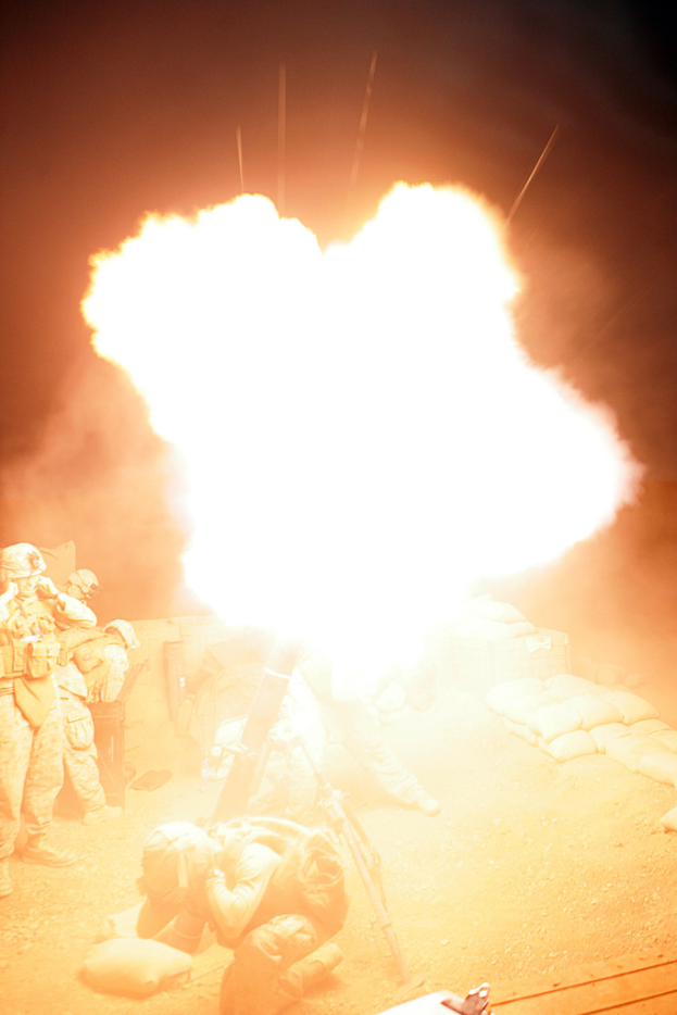 Mehrere Personen in Uniformen und Helmen befinden sich auf einem Gelände mit einigen Taschen, wobei in der Mitte des Bildes ein Feuer brennt.