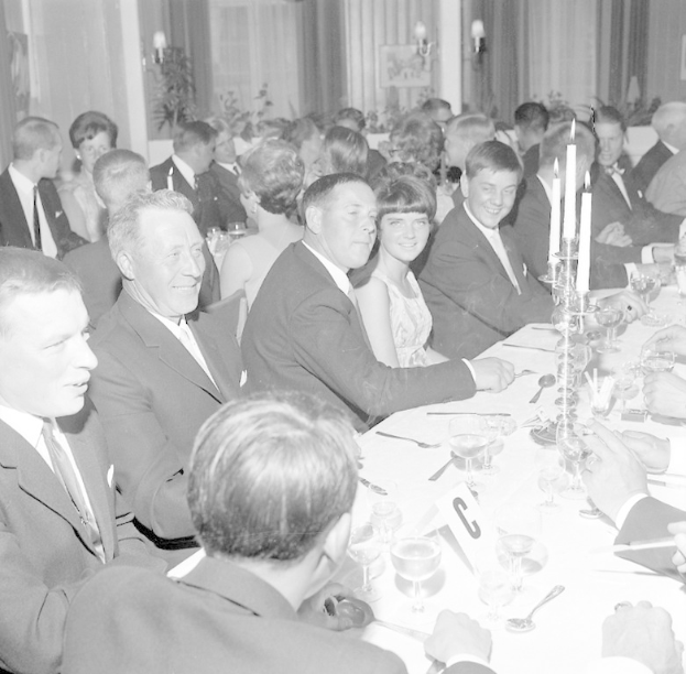 Eine Gruppe von Menschen sitzt um einen langen Tisch, lächelt und genießt die Gesellschaft des anderen, mit Gläsern, Besteck, Kerzen und anderen Gegenständen auf dem Tisch und Vorhängen und Wandlampen im Hintergrund.