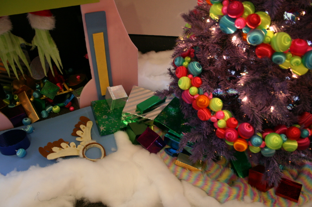 Drinnen steht rechts ein geschmückter Weihnachtsbaum mit Lichterketten und Schmuck, in der Mitte Geschenkboxen und Dekorationen, links oben eine Hütte mit Dekorationen und unten Wolle und Stoff.