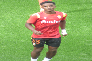 Emmanuel Adebayor, ein Mann in einem roten T-Shirt und schwarzen Shorts, geht auf einem Fußballfeld mit zwei Personen im Hintergrund.