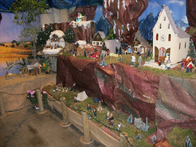 Eine Krippe mit einem kleinen Dorf in der Mitte, umgeben von Häusern, Bäumen, Pflanzen, Blumen, Gras, Steinen und anderen Gegenständen, mit einem Vorhang oben, der die festliche Stimmung unterstreicht.