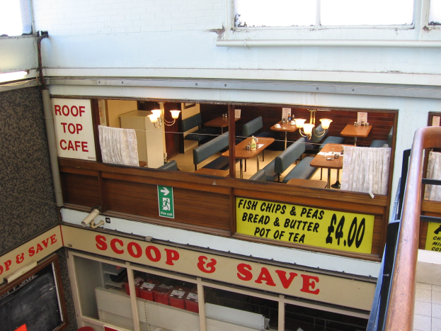 Ein cafeartiges Restaurant mit Tischen, Stühlen und beleuchteten Tafeln mit Text, das eine Brüstung auf der rechten Seite und eine Wand im Hintergrund mit einer 'Scoop & Save'-Aufschrift hat.