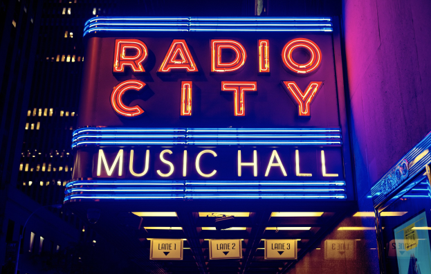 Das erleuchtete Radio City Music Hall in New York City, umgeben von hohen Gebäuden, mit einer 'Radio City'-Leuchtschrift und zusätzlichen Schildern rechts.