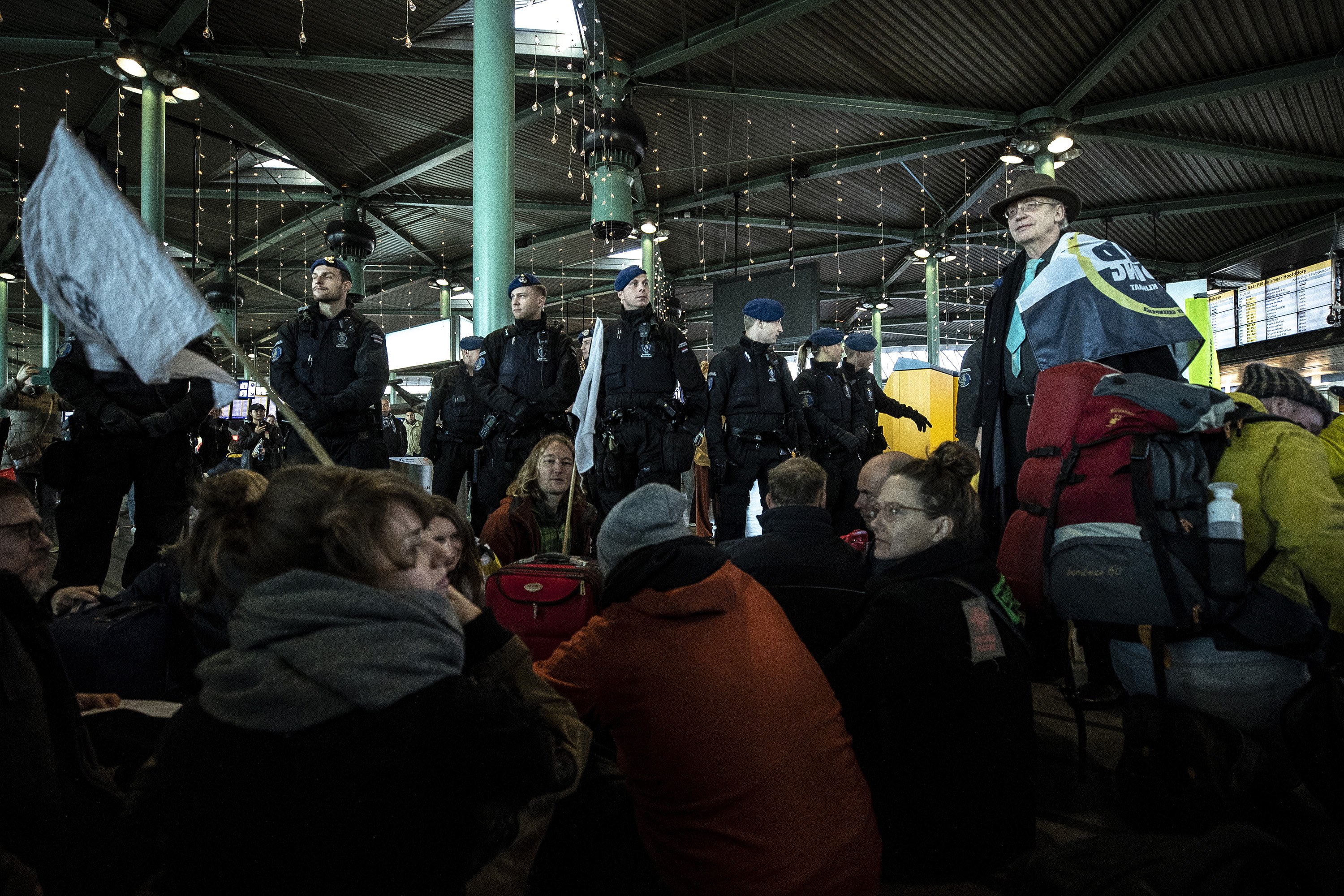 Eine Gruppe von Menschen steht vor einer Menge in einem Bahnhof, wobei einige Personen Schilder und Transparente halten, was auf eine Demonstration hindeutet.