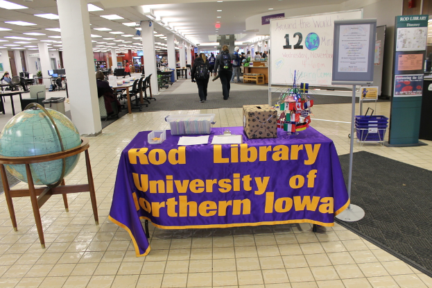 Eine Ausstellung mit einem Tisch, der verschiedene Landesflaggen, eine Box, Papiere und ein Banner zeigt, eine Kugel links daneben, eine Anschlagtafel rechts mit ein paar Menschen in der Nähe und mehreren Tischen und Stühlen links.