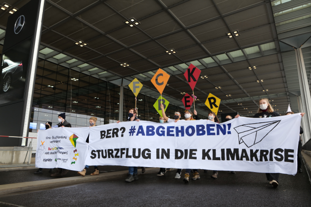Eine Gruppe von Menschen mit Masken hält ein Banner mit der Aufschrift "Mercedes-Benz Sturzflug in die Klimakrise" vor einer Mercedes-Benz-Ausstellung mit Glaswänden und Deckenbeleuchtung, mit einem Logo auf der linken Seite.