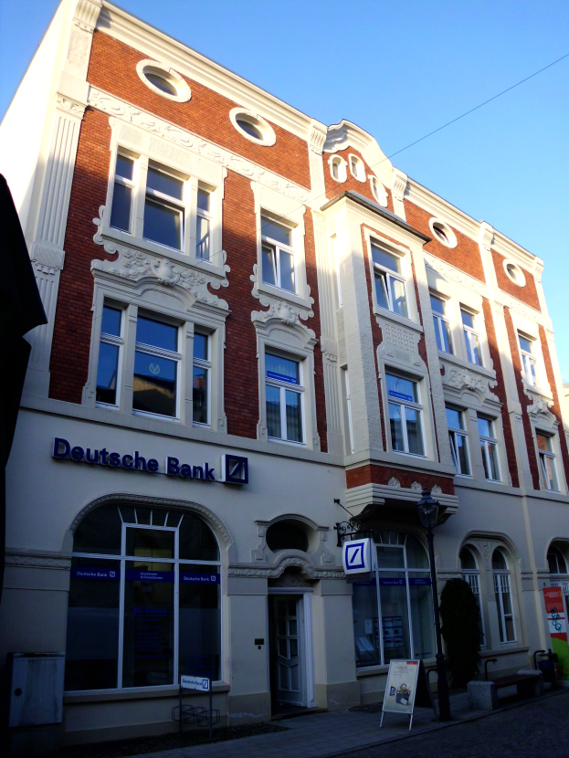 Ein Gebäude mit einem "Deutsche Bank"-Schild an seiner Seite, umgeben von Straßenmasten, Straßenlaternen, Namenschildern, Büschen und einem klaren blauen Himmel.