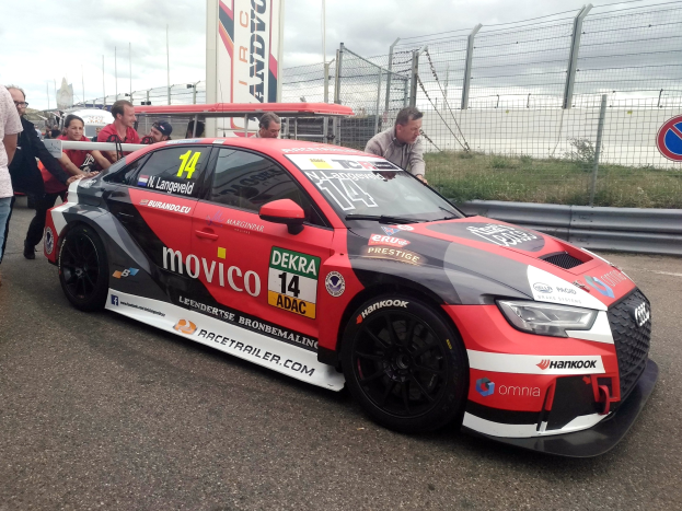 Audi RS3 DTM-Rennwagen auf einer Straße mit einer Gruppe von Menschen im Hintergrund, umgeben von einem Zaun, Gras, einem Schild, einer Fahne mit Text, Pfosten und einem bewölkten Himmel.