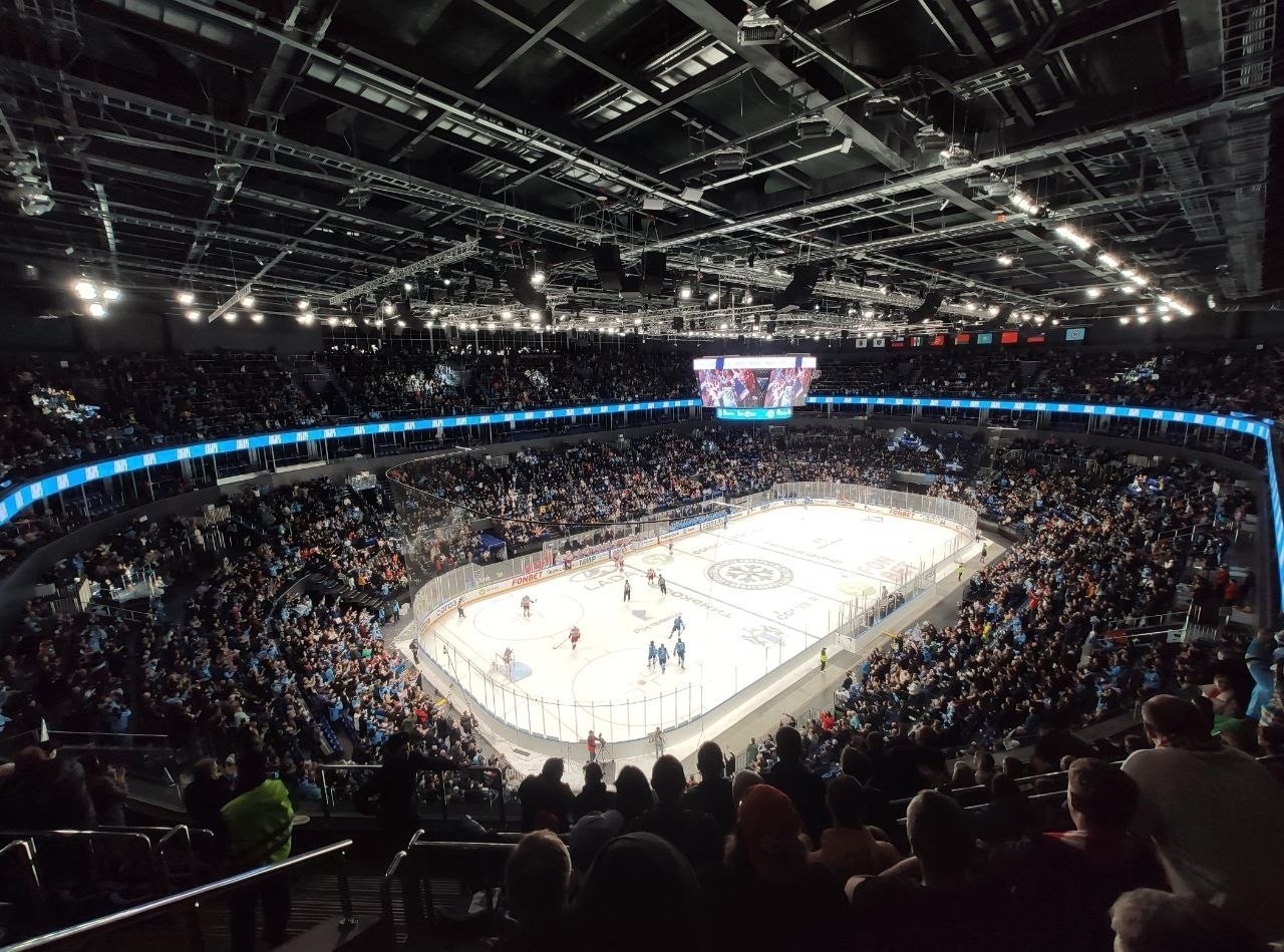 Eishockeyspiel in einer großen Arena mit Zuschauern auf den Rängen und Personal am Boden, mit Geländern, Überwachungsbildschirmen und heller Beleuchtung, identifiziert als O2 Arena in London.