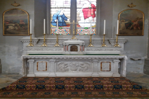 Innere einer Kirche mit großem Buntglasfenster, gerahmten Fotos an den Wänden, Kerzenständern, einem Teppichboden und kunstvoll verzierten Wänden.