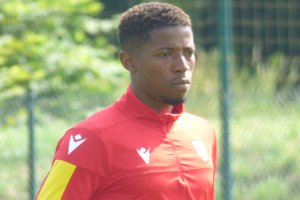 Ein junger Mann in einem roten und gelben T-Shirt steht auf einem Fußballfeld umgeben von einem Zaun und Bäumen und sieht entschlossen aus.