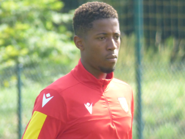 Ein junger Mann in einem roten und gelben T-Shirt steht auf einem Fußballfeld umgeben von einem Zaun und Bäumen und sieht entschlossen aus.