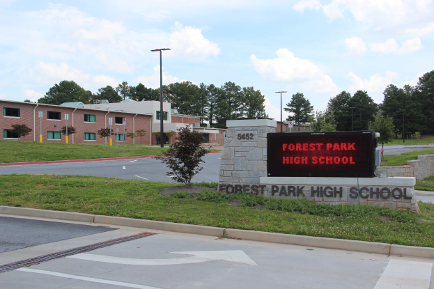 Außenansicht der Forest Park High School mit Straße, Gras, textbedruckter Wand, Laternen, Bäumen, Gebäuden mit Fenstern und bewölktem Himmel.