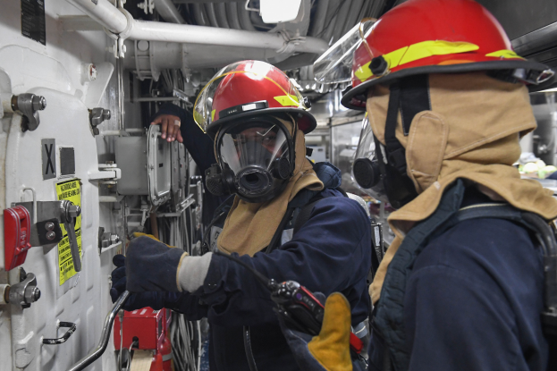 Zwei Feuerwehrleute in Schutzausrüstung, einschließlich Helmen und Gasmasken, arbeiten an einem Boot, mit einer Wand mit verschiedenen Gegenständen auf der linken Seite und Rohren und anderen Gegenständen im Hintergrund.
