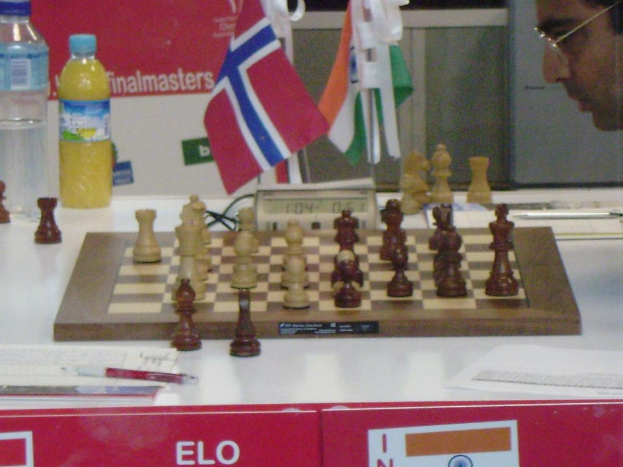 Ein Schachbrett, Timer, Flasche, Buch und Stift sind auf einem Tisch mit einer Person mit Brille im Vordergrund und Flaggen an der Wand im Hintergrund.