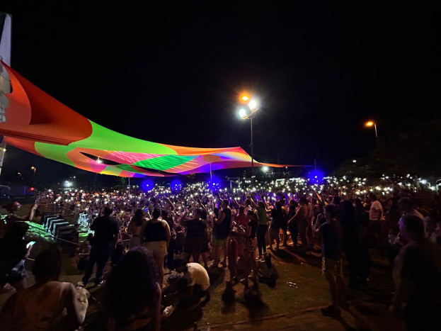 Eine große Menschenmenge steht vor einem hell erleuchteten Zelt in der Nacht während des 2018er Kunstfestivals, mit farbigen Lichtern und Pfählen, die die Szene beleuchten, einem Gebäude auf der linken Seite und dem Nachthimmel im Hintergrund.