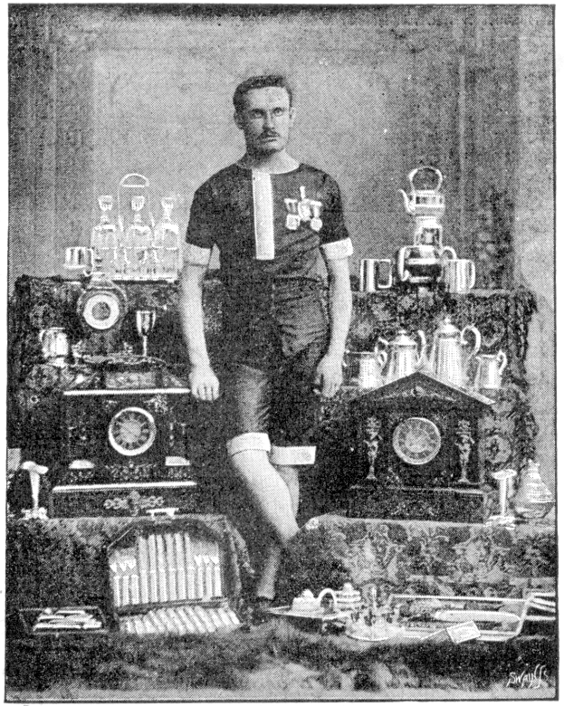 Ein Schwarz-Weiß-Foto eines Mannes in einem Fußballtrikot, der vor einem Tisch voller Trophäen und Pokale steht, mit der Aufschrift '1909-1910 WM-Sieger' unten.