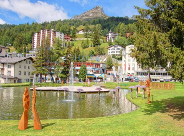 Ein zentraler Wasserplatz mit Brunnen, umgeben von Gebäuden, Bäumen und Bergen, mit Gras am Boden des Bildes.