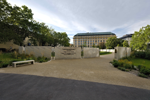 Eine Parkbank umgeben von Bäumen, Pflanzen und Gras, mit Gebäuden und einem bewölkten Himmel im Hintergrund und Text an der Wand hinter der Bank, wahrscheinlich in Zusammenhang mit dem Holocaust-Mahnmal in Berlin.