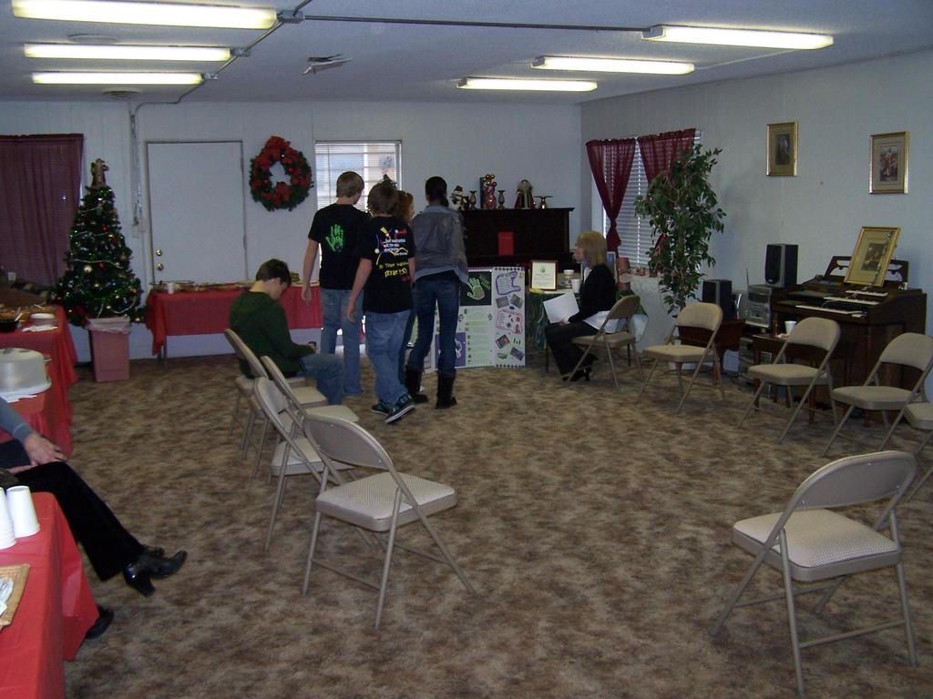 Ein Raum mit mehreren Stühlen, Tischen und Menschen, einige stehen, andere sitzen. Auf den Tischen stehen Becher und Gefäße. Im Hintergrund ist eine Wand mit einem Blumenstrauß, einem Vorhang, einer Tür, Fotorahmen und Fenstern zu sehen. Ein Weihnachtsbaum und ein Piano sind vorhanden, mit Röhrenleuchten an der Decke.