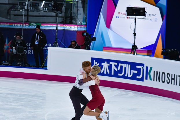 Ein Paar umarmt sich beim Eislaufen auf einer Eisbahn, im Hintergrund stehen ein paar Menschen, einige halten Kameras, und eine Tafel mit Schrift darauf.