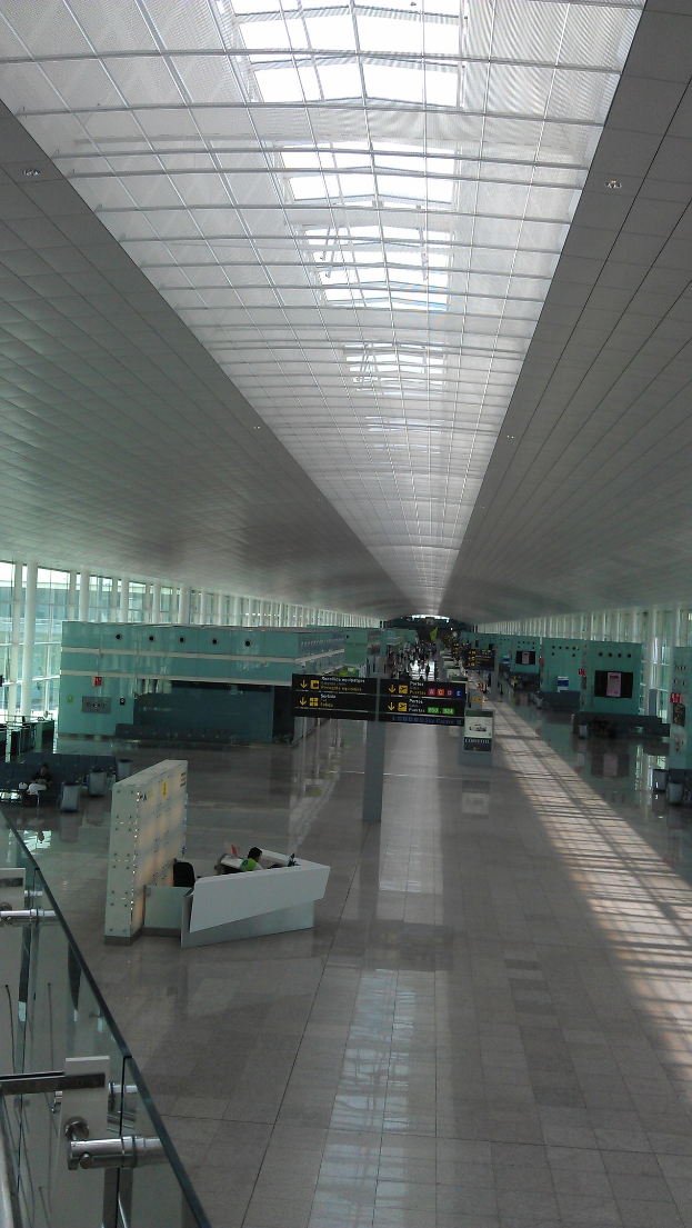 Innenansicht eines Flughafen-Terminals mit einer Glasbrüstung auf der linken Seite, einem Boden darunter, einer Decke mit Lampen darüber und mehreren Menschen im Hintergrund.