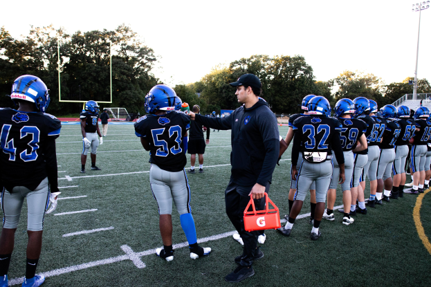 Football coach in a cap speaking to players in helmets on a field with goal post, trees, poles, lights, railings, and a clear blue sky.